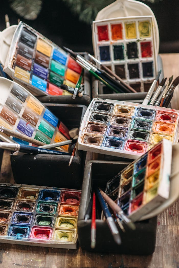 our-services-3 A collection of colorful watercolor paint sets and brushes arranged on a wooden table.